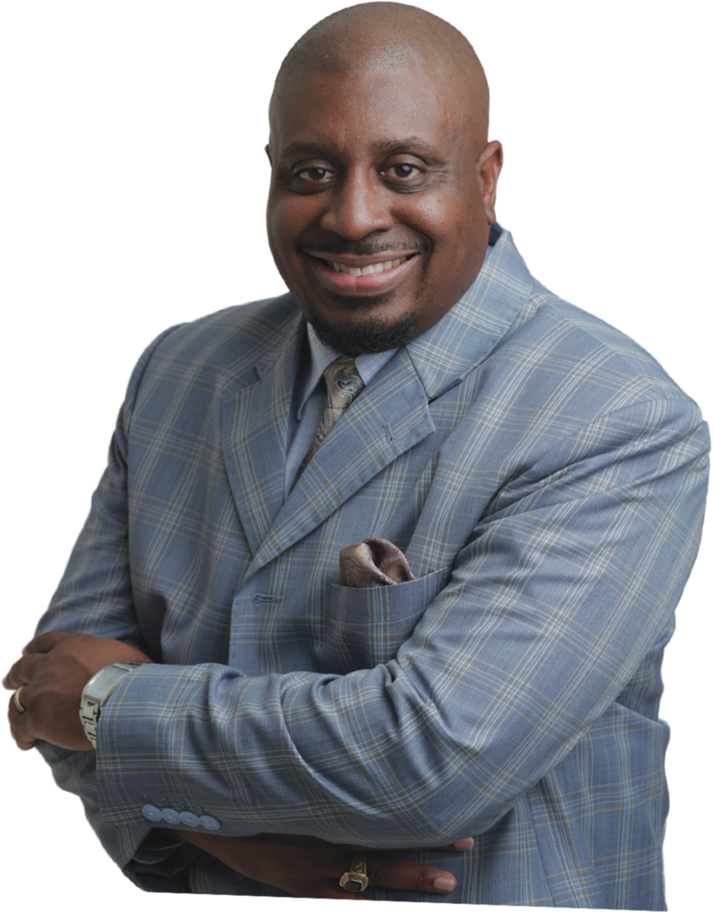 A professional studio portrait of a Black man with a goatee smiling warmly with his arms crossed against a solid black background. He is wearing a light blue and grey plaid suit jacket, a collared shirt, a gold patterned tie, and a matching pocket square.