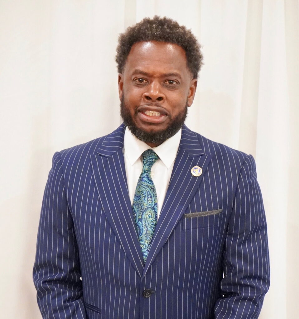 A professional studio portrait of a Black man with a well-groomed beard looking directly at the camera against a white background. He is wearing a navy blue pinstriped suit jacket, a white collared dress shirt, and a vibrant blue and green paisley tie.