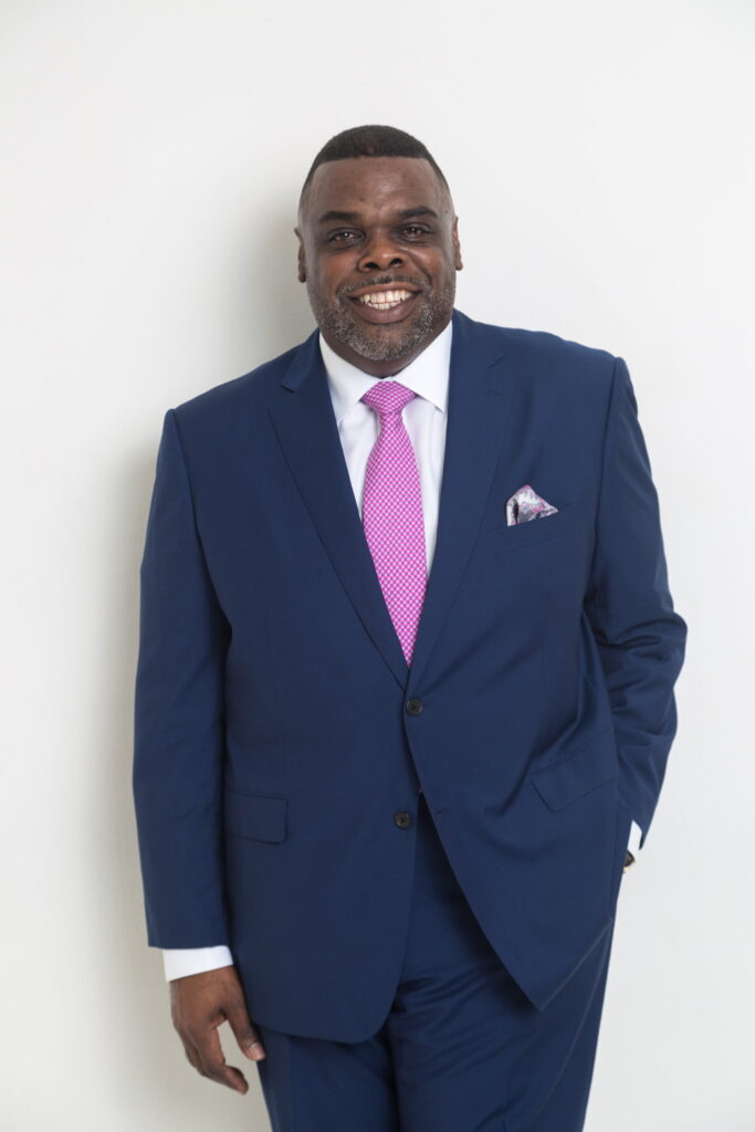A professional studio portrait of a Black man smiling brightly with one hand in his pocket against a white background. He is wearing a tailored royal blue suit, a white dress shirt, a solid pink tie, and a multi-colored patterned pocket square.