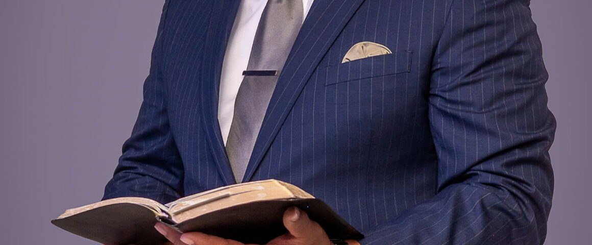 A professional studio portrait of a bald Black man with a grey-speckled beard, standing against a solid grey background. He is dressed in a dark blue pinstriped suit with a grey tie and is holding an open Bible, looking forward with a serene expression.