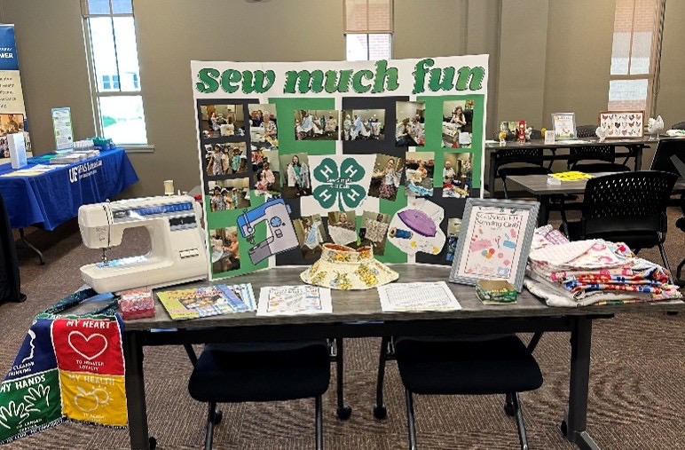 A 4-H club exhibition table inside a classroom or community room. The table features a trifold display board titled 'sew much fun' with the 4-H clover logo and photos of kids sewing. On the table sits a white Brother electric sewing machine, a handmade floral visor, informational pamphlets, a framed sign, and a stack of colorful patterned quilts.