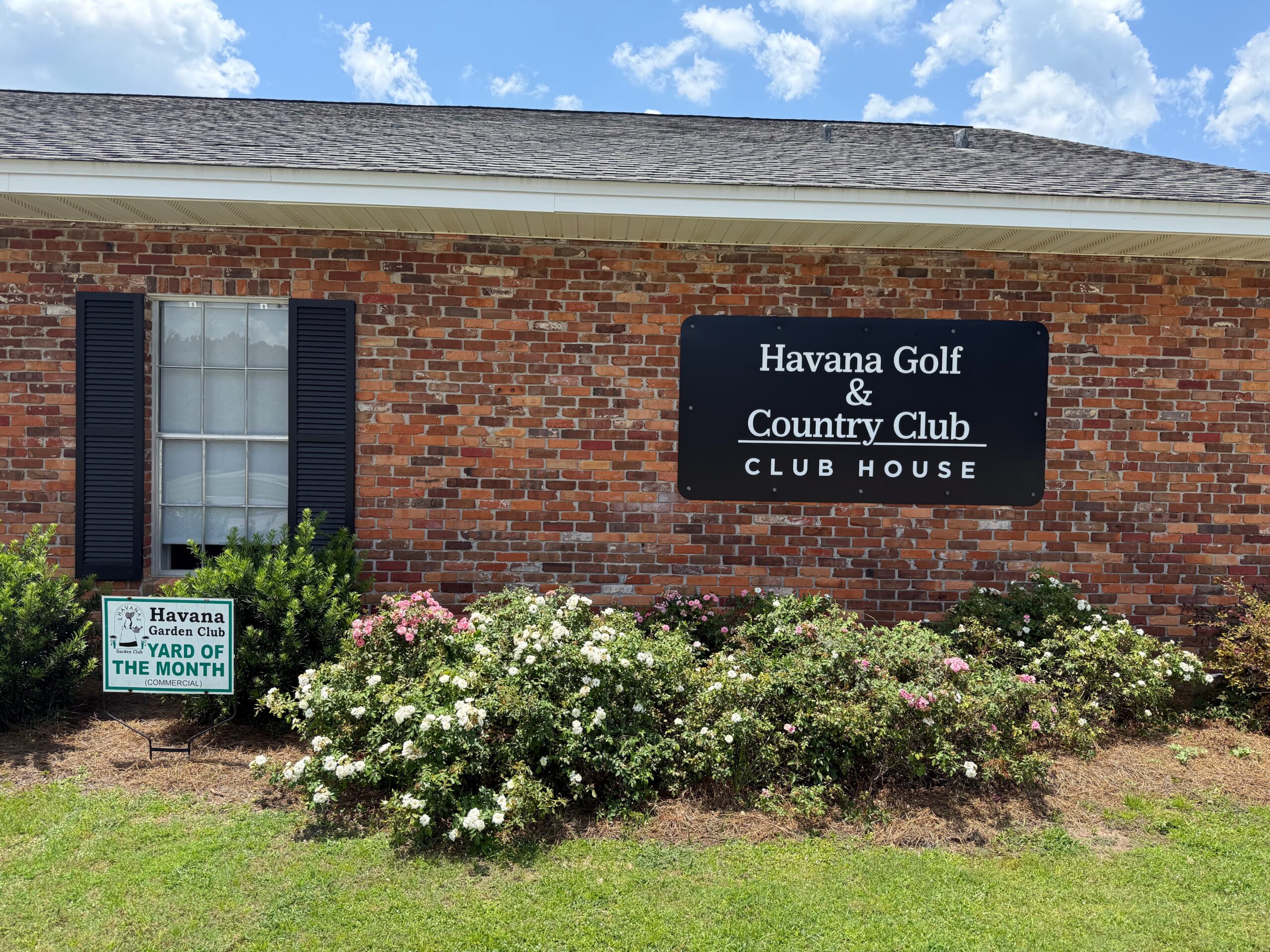 The exterior red brick wall of the Havana Golf & Country Club Clubhouse. A black sign on the wall reads 'Havana Golf & Country Club CLUB HOUSE' in white text above a bed of pink and white flowering bushes. On the left, a small green sign reads 'Havana Garden Club YARD OF THE MONTH'.