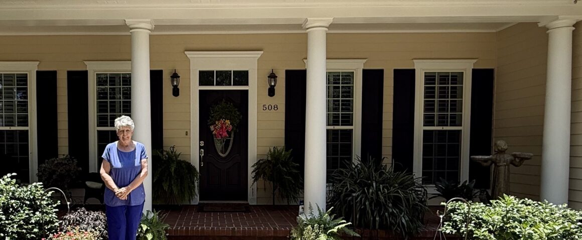 An elderly woman with short white hair standing on the front sidewalk of a beige single-story house with white columns and dark shutters. She is wearing a blue top and blue pants, surrounded by potted plants, hanging flower baskets, and lush green landscaping.