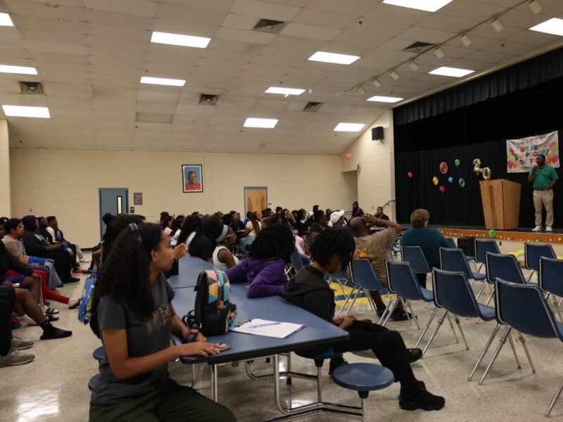 A large group of students seated at cafeteria tables and in rows of chairs in a school auditorium, listening to a man speaking at a wooden podium on a stage. The stage features a black backdrop decorated with colorful paper fans, gold balloons spelling '20', and a partially visible 'We Are Proud of You' banner.