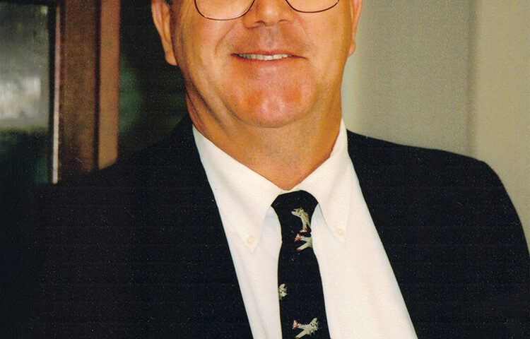 Portrait of a middle-aged man with short gray hair and wire-rimmed glasses smiling at the camera. He is wearing a dark suit jacket, a white collared shirt, and a dark tie with a pattern of small airplanes.