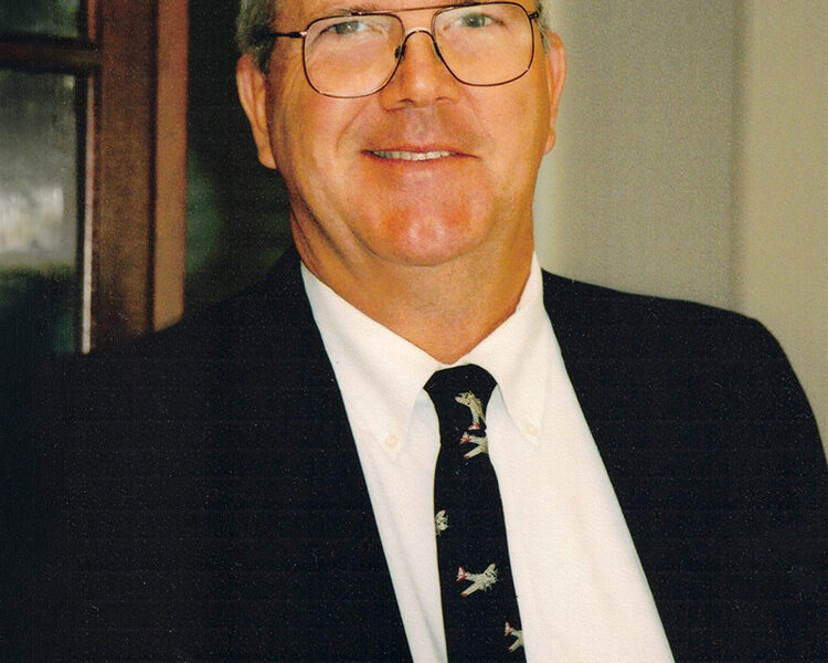 Portrait of a middle-aged man with short gray hair and wire-rimmed glasses smiling at the camera. He is wearing a dark suit jacket, a white collared shirt, and a dark tie with a pattern of small airplanes.