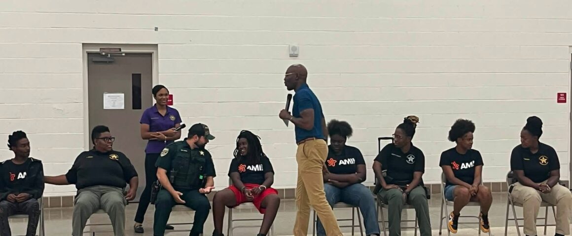 A man standing in front of a group of seated teenagers and police officers in a gymnasium, holding a microphone and addressing the audience.