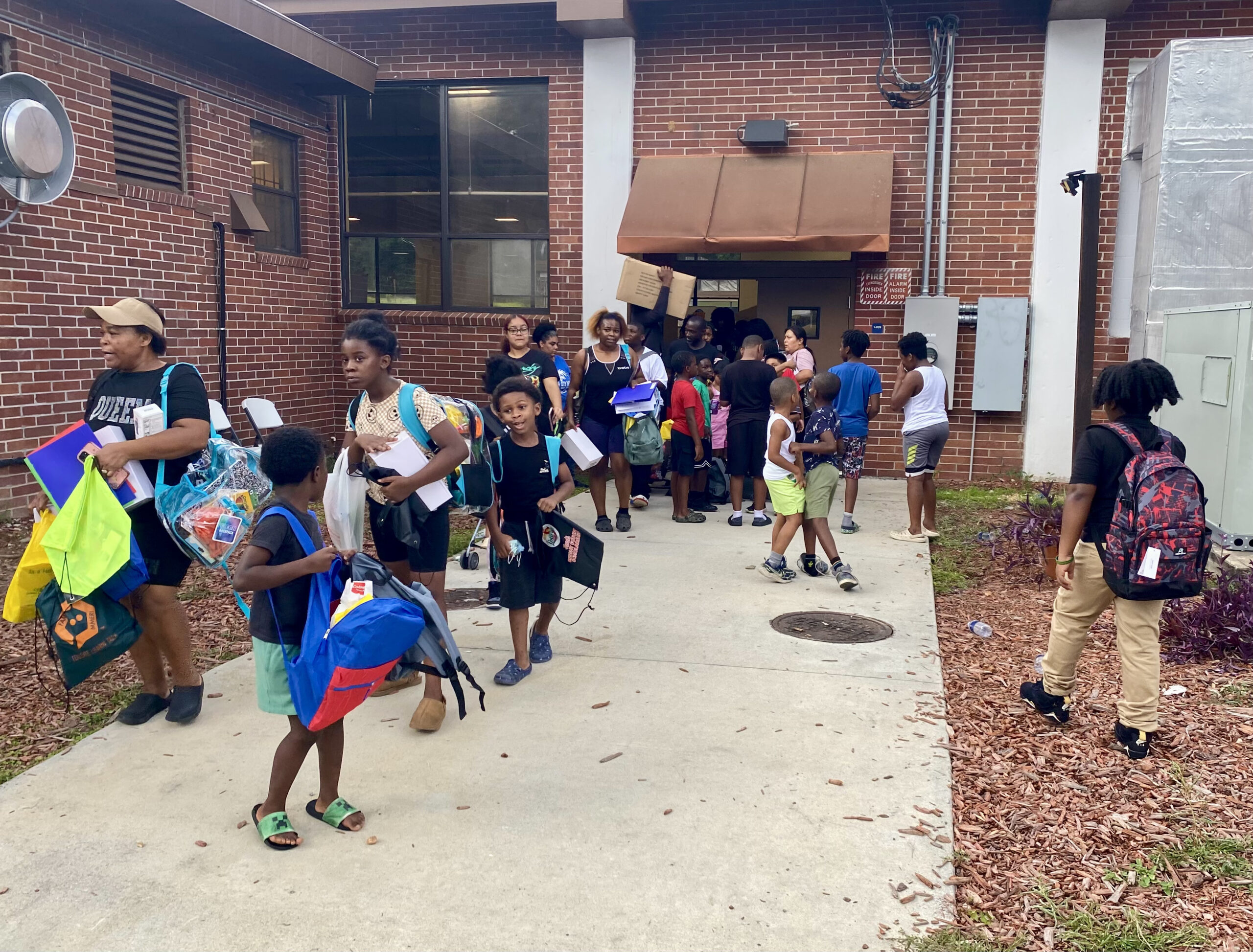 Children and parents carrying new backpacks, folders, and school supplies walking out of a brick building door along a concrete walkway.