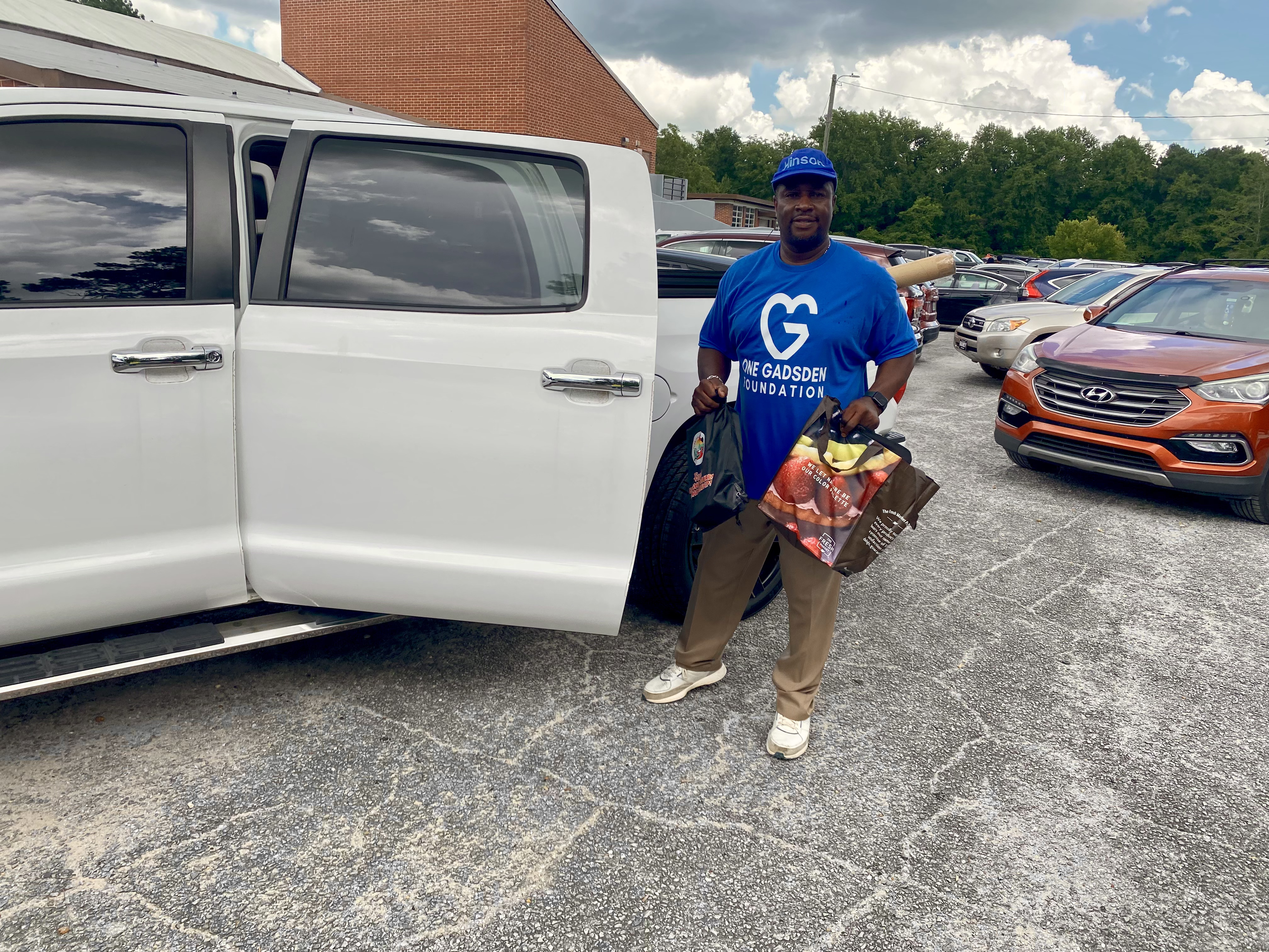 A man wearing a blue 'One Gadsden Foundation' t-shirt and matching hat standing next to an open white pickup truck door, holding two filled tote bags.