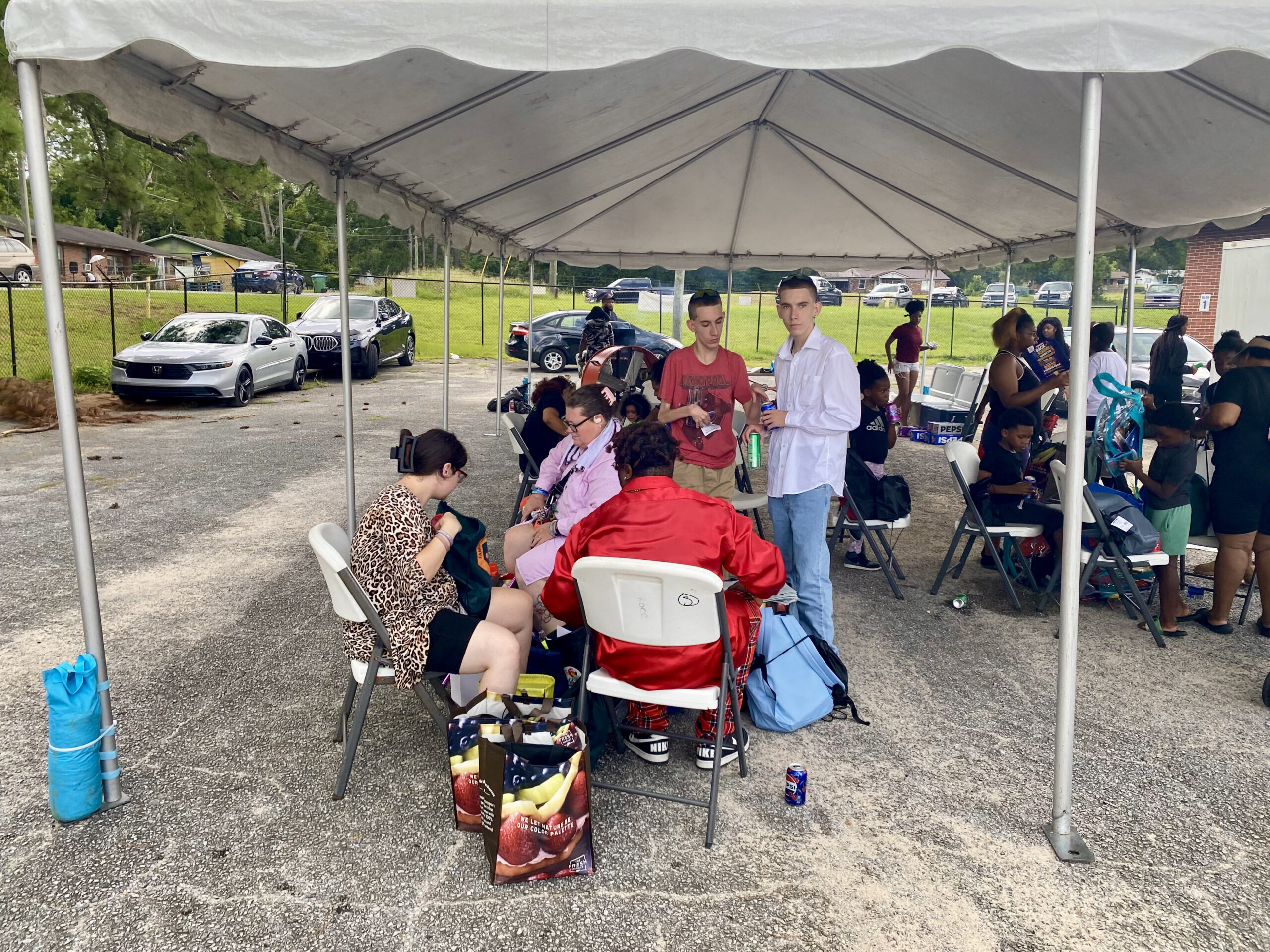 A group of people sitting on folding chairs underneath a large white event tent setup outdoors in a paved parking lot area, sorting through bags and supplies.