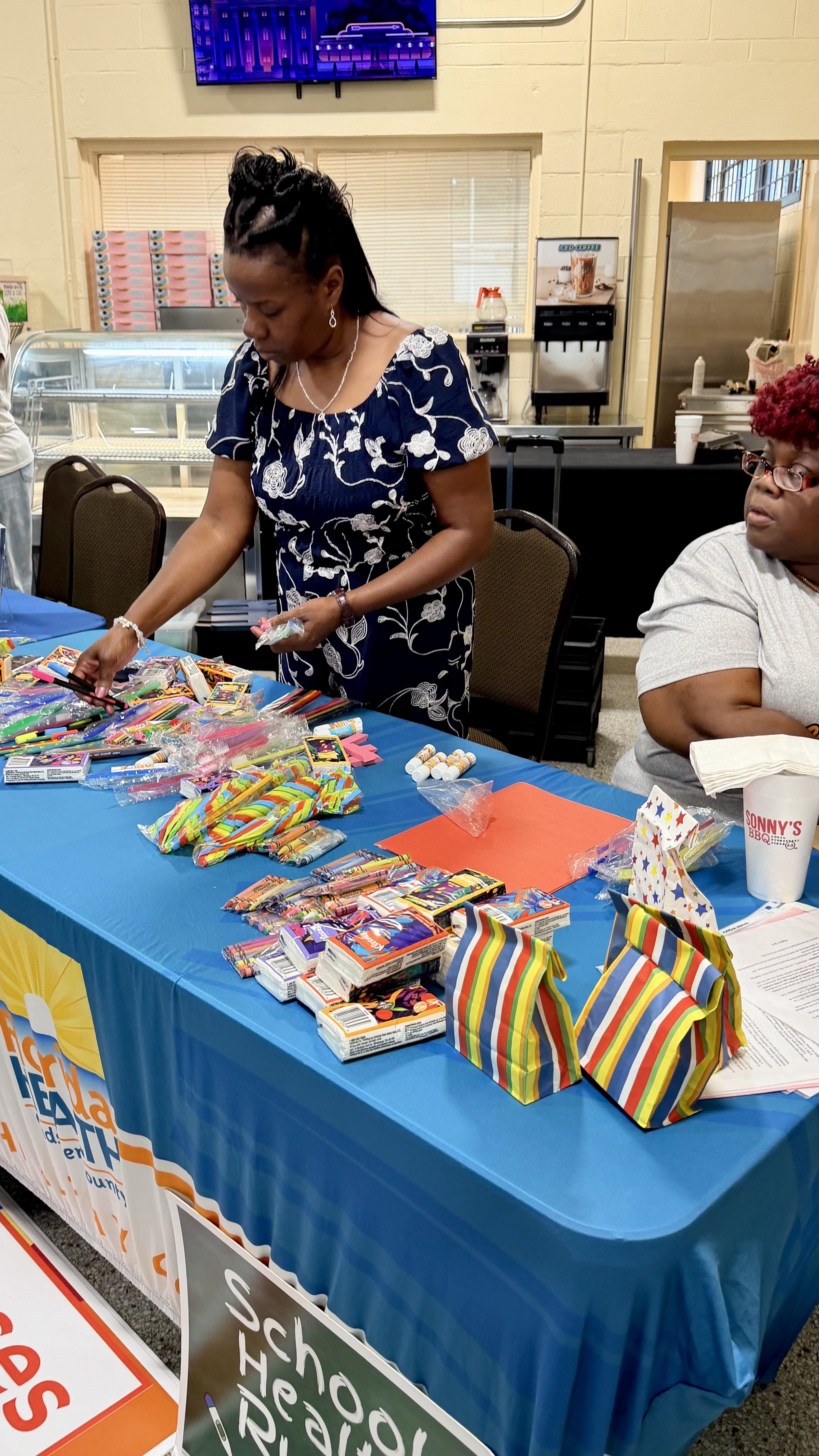 A woman in a navy floral dress organizing school supplies including markers, crayons, and small gift bags laid out on a table draped with a blue tablecloth.