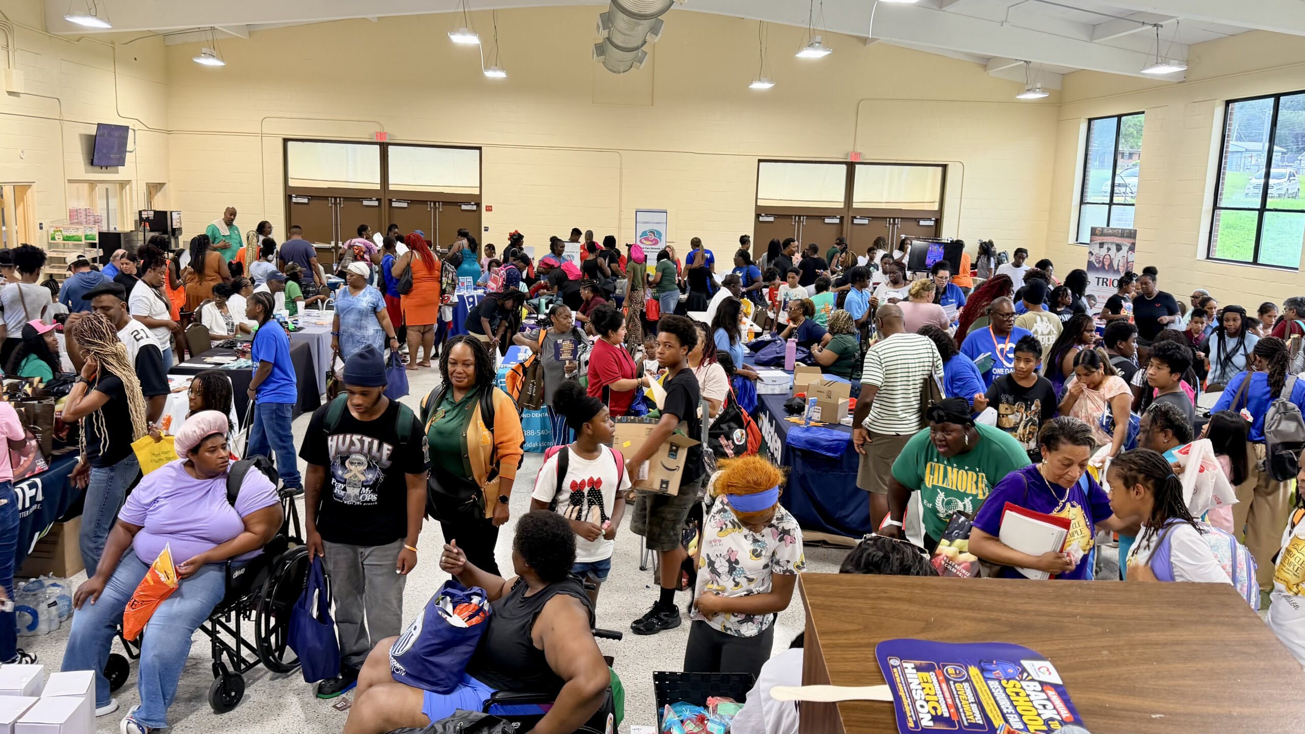 An aerial wide-angle view of a crowded indoor gymnasium filled with parents, children, and vendors attending a community back-to-school resource fair.