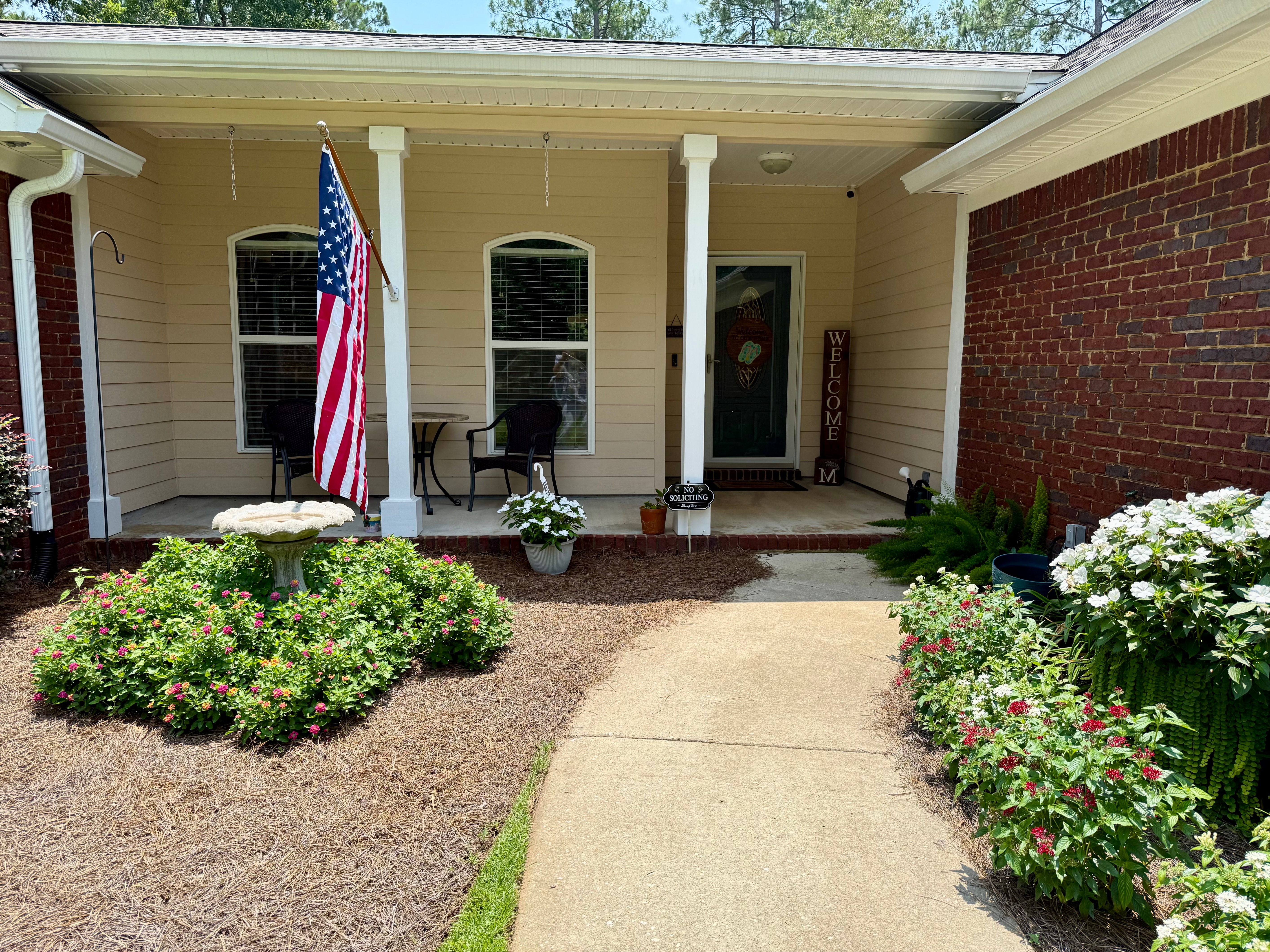 A concrete walkway leading to a covered home front porch with light yellow siding, white columns, an American flag, a bird bath surrounded by colorful flowers, potted plants, and two patio chairs.