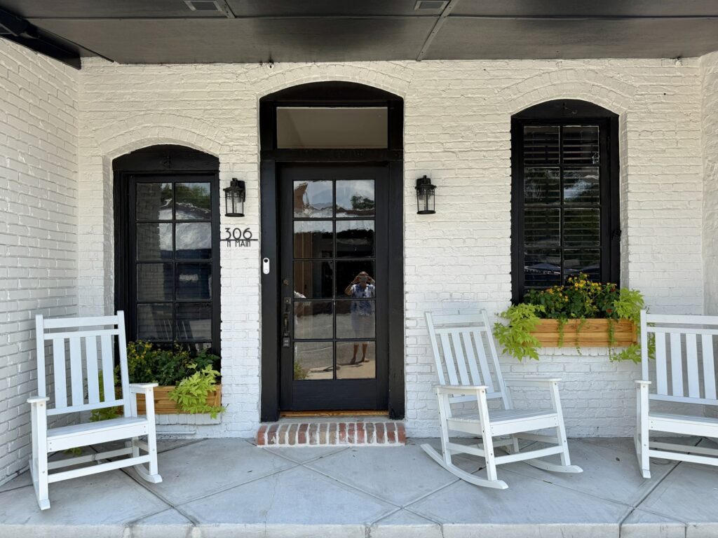 The porch of a white painted brick building featuring three white wooden rocking chairs, two windows with wooden planter boxes filled with yellow and green plants, and a dark glass panel front door labeled 306 N Main.