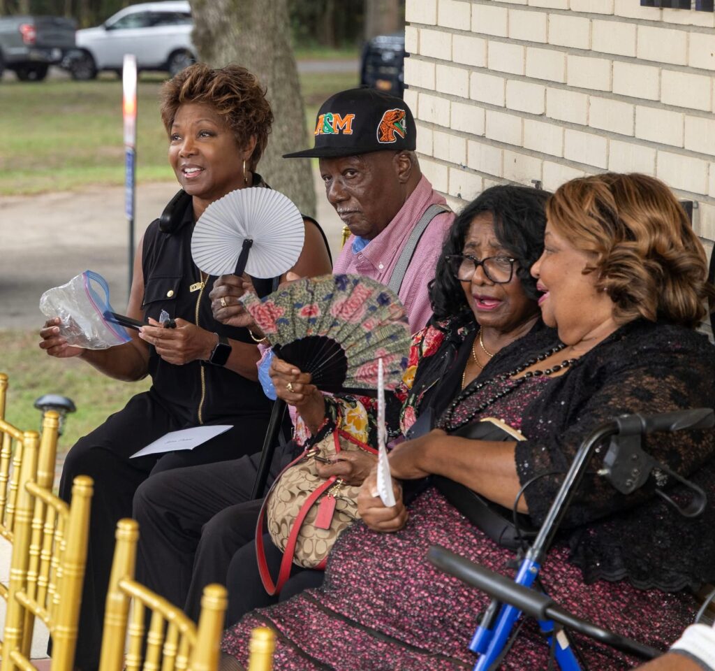 Four community members seated outdoors on gold chairs during an event, cooling themselves with hand fans.