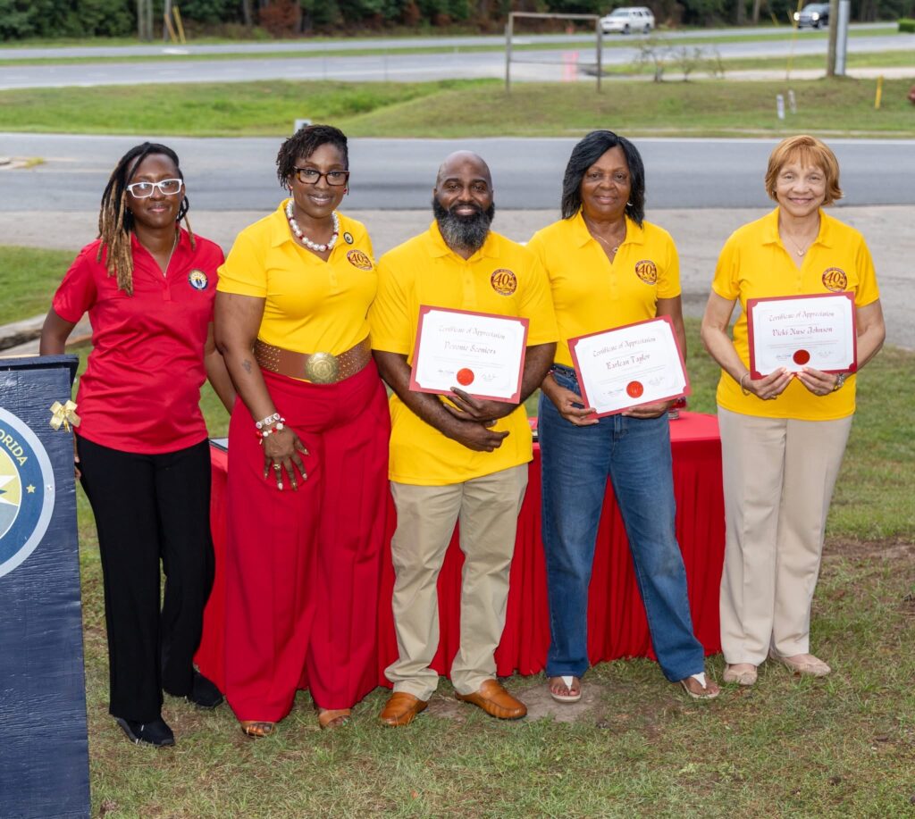 Five community members wearing matching red and yellow polo shirts holding certificates of appreciation at an outdoor anniversary event