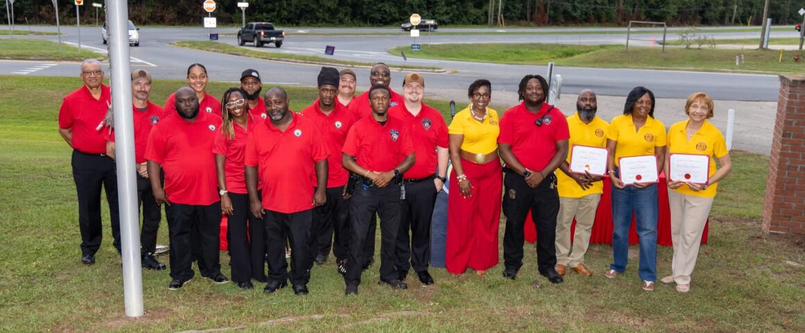 A large group pose of city staff, officials, and committee members wearing red and yellow shirts standing outdoors.