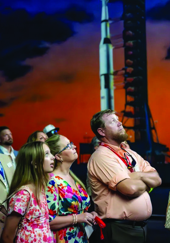 Roger and Bridgette Rankin with their daughter Lilly looking up while standing in front of a Saturn V rocket exhibit backdrop during the state conference.