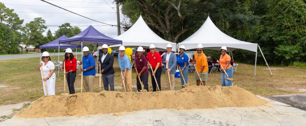 Local officials and project leaders in hard hats breaking ground with shovels at a dirt pile during an outdoor groundbreaking event under canopy tents.