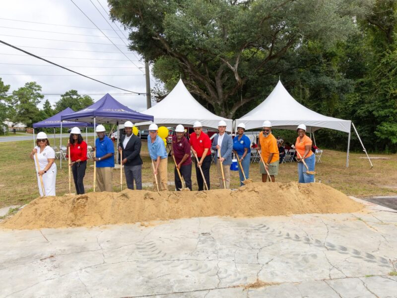 Local officials and project leaders in hard hats breaking ground with shovels at a dirt pile during an outdoor groundbreaking event under canopy tents.