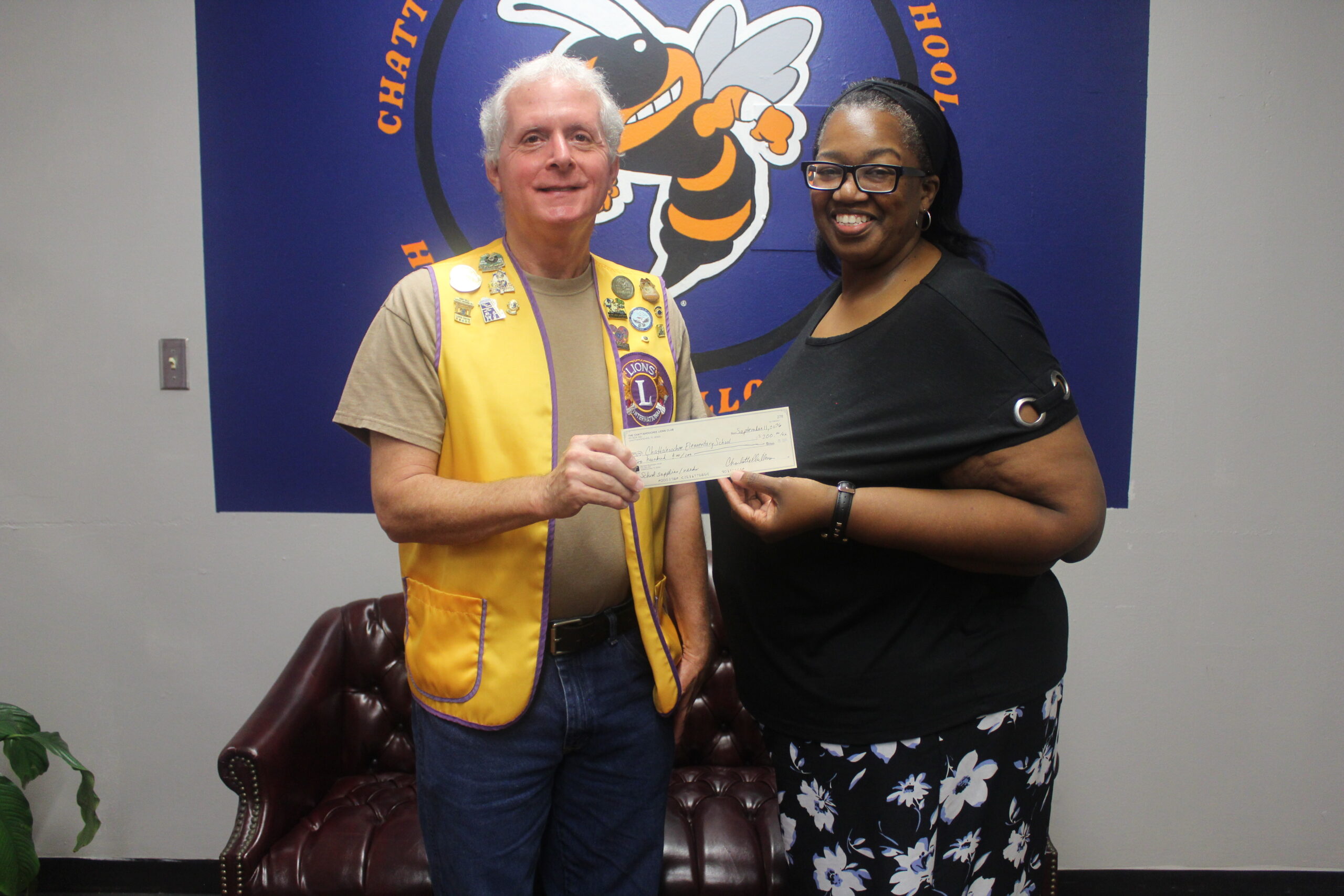 A Lions Club representative in a yellow vest posing alongside a woman while co-holding a donation check in front of a Chattahoochee school wall logo.