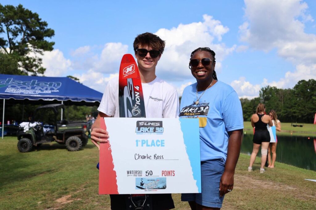 A young man holding a water ski and a 1st place award poster alongside a woman wearing a blue event shirt and sunglasses in front of a lake.