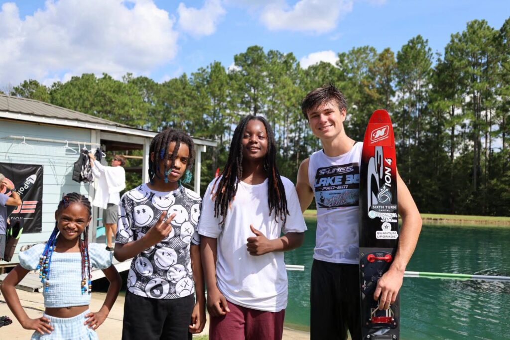 A group of young people and a water skier holding a red and black slalom ski posing together outdoors by the edge of Lake 38.
