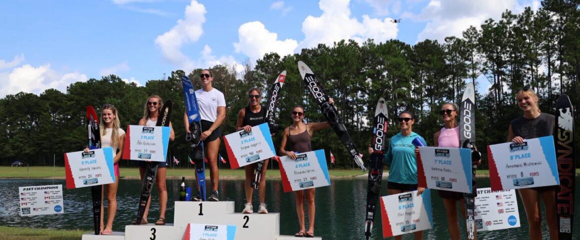 Top eight female water ski competitors standing on a three-tiered podium holding placement signs and skis at an outdoor lake venue.