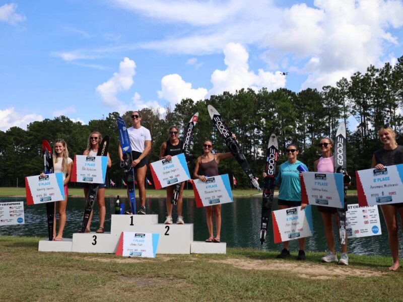 Top eight female water ski competitors standing on a three-tiered podium holding placement signs and skis at an outdoor lake venue.
