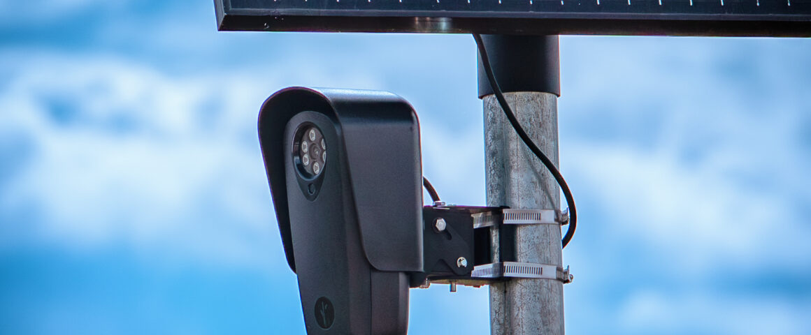 A close-up photograph of a black Flock Safety license plate reader camera and solar panel mounted on a metal pole against a blue sky.