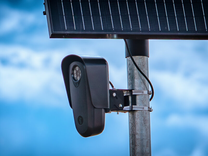A close-up photograph of a black Flock Safety license plate reader camera and solar panel mounted on a metal pole against a blue sky.