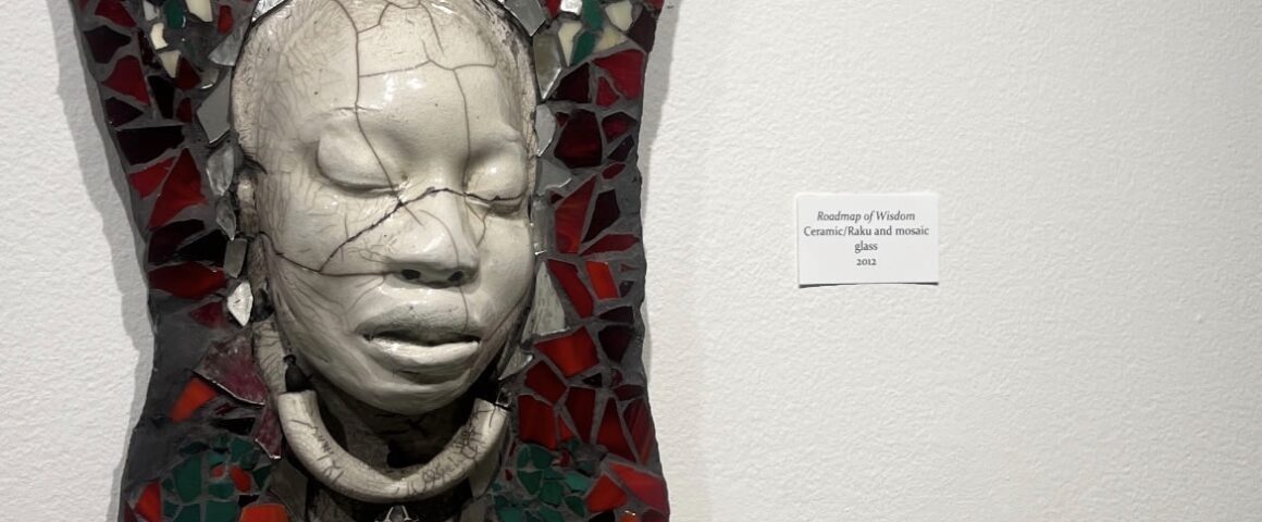 A ceramic and glass mosaic artwork titled 'Roadmap of Wisdom' featuring a white face mask against red and green glass pieces mounted on a wall.