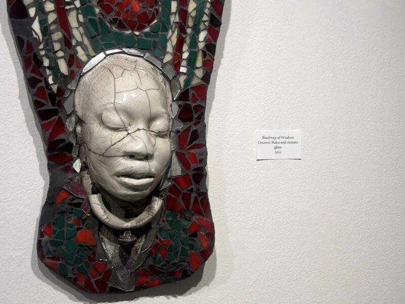 A ceramic and glass mosaic artwork titled 'Roadmap of Wisdom' featuring a white face mask against red and green glass pieces mounted on a wall.
