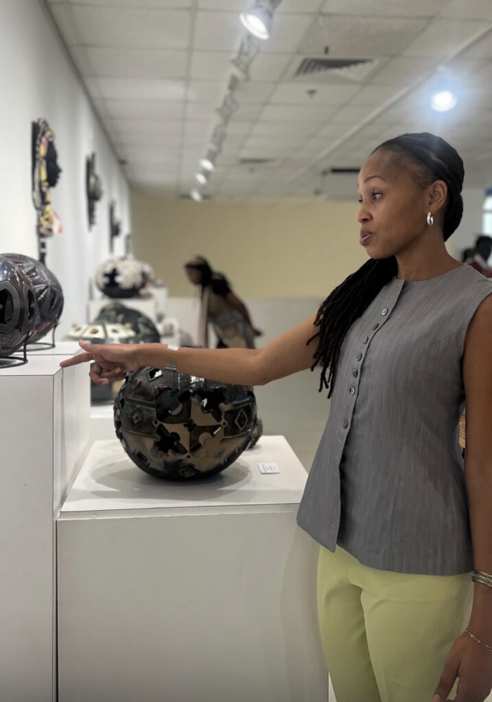 A woman with braided hair wearing a grey sleeveless button-up top pointing at ceramic orb sculptures on display pedestals inside an art gallery.