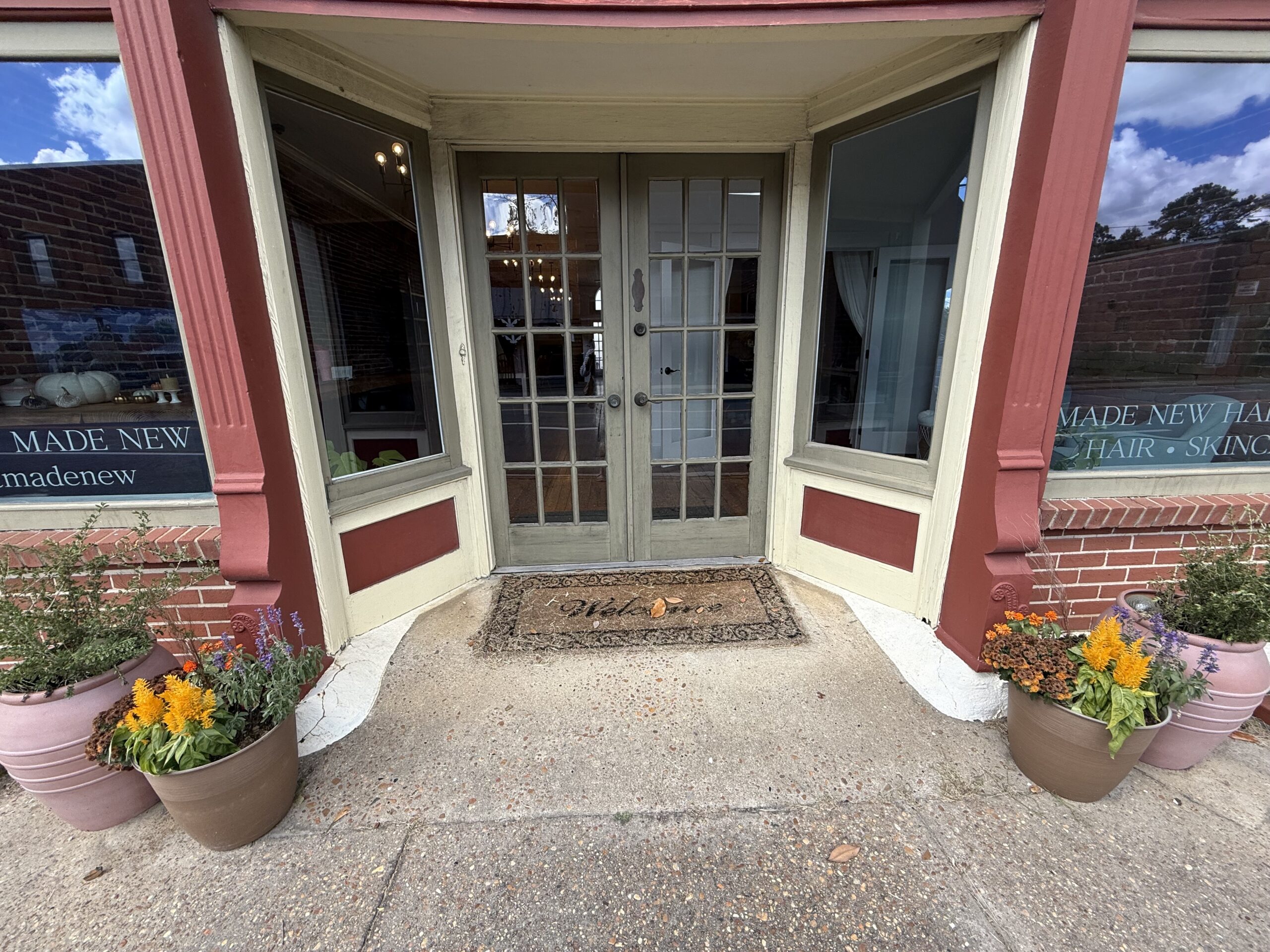 The glass double-door entrance of Made New Hair & Skincare salon framed by red brick pillars and potted flowers.