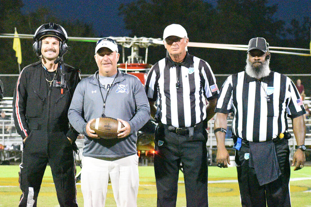A football coach holding a ball flanked by two referees and a helicopter pilot standing on a field before a game.