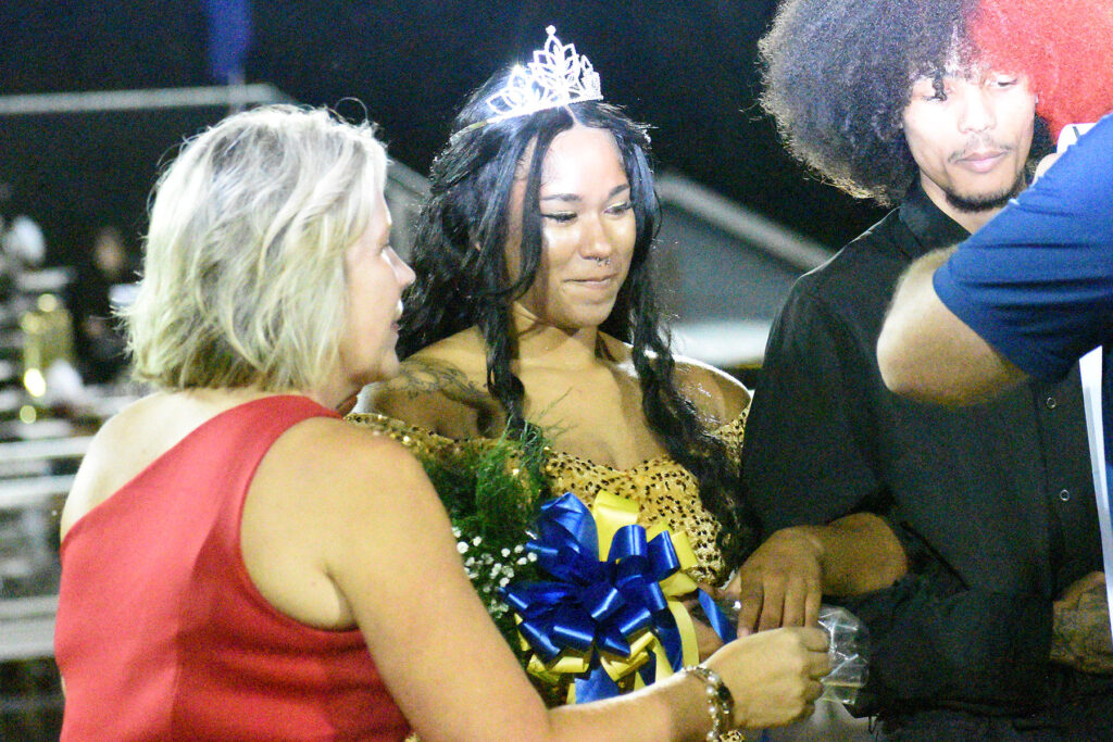 A young woman wearing a crown and a gold dress receiving flowers and congratulations at a homecoming ceremony.