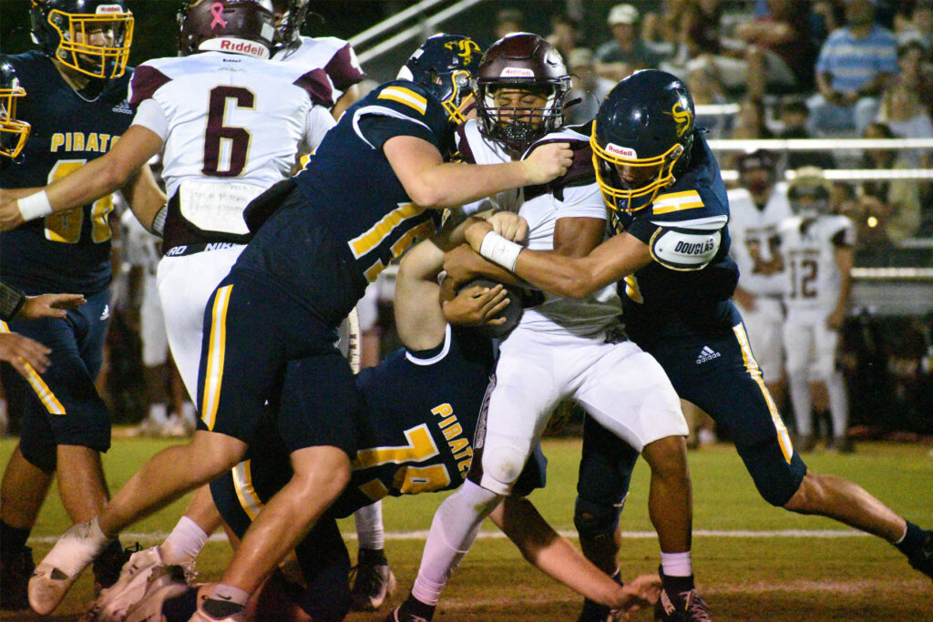 Action shot of high school football players tackling a carrier during a game under stadium lights.