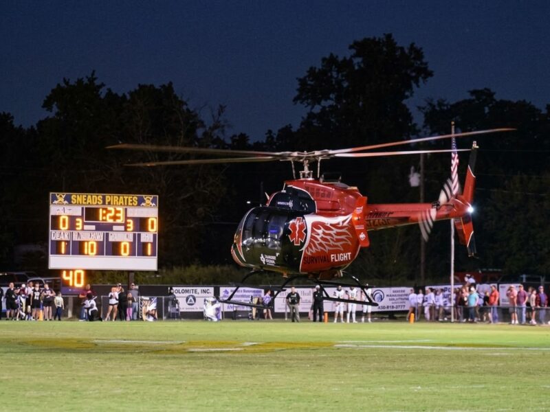 A red Survival Flight medical helicopter hovering above a football field next to a scoreboard showing the Sneads Pirates.